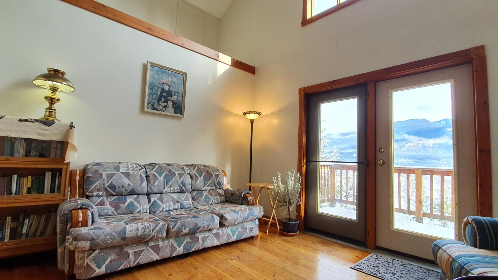 Lounge area with sofa, chairs and bookstand. A double glazed door leads to an exterior balcony with wooden railing and mountain view.